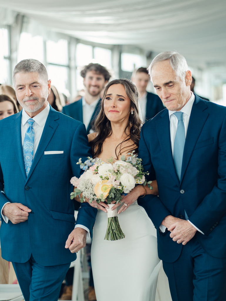 bride getting emotional walking down the aisle with her dad and stepdad during her wedding ceremony at Silver Swan Bayside
